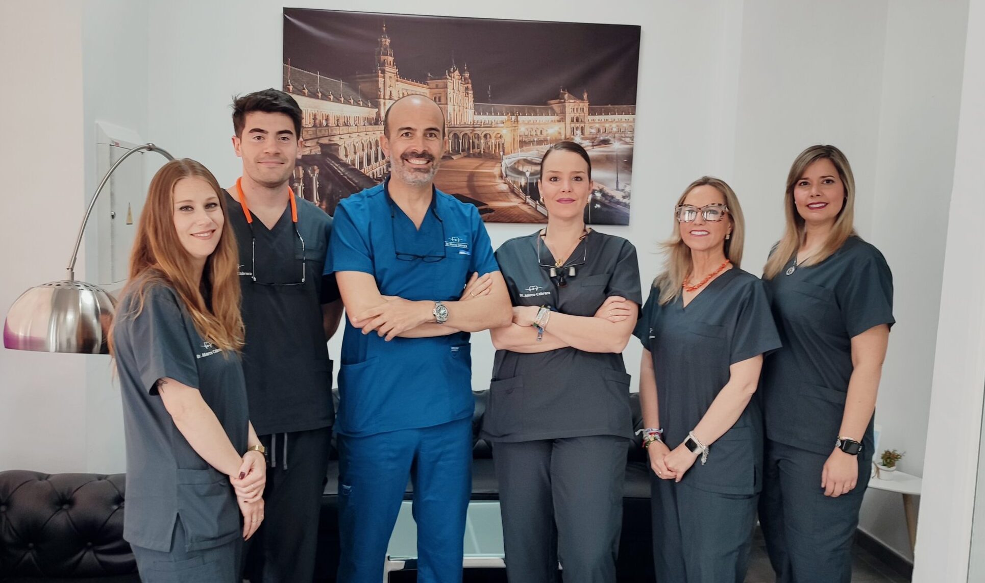Foto de grupo del equipo completo de profesionales de la Clínica Dental Dr. Marco Cabrera en Alcalá del Río. Siete miembros, incluyendo dentistas e higienistas, sonriendo en la recepción moderna, dedicados a la salud bucodental de los pacientes.
